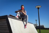 Mujer caucásica con mascarilla facial, sentada en la pared con monopatín y usando smartphone. pasando el rato en skatepark urbano en verano durante coronavirus covid 19 pandemia. - foto de stock