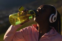 Donna afroamericana che beve acqua, si prende una pausa all'aperto, indossa le cuffie. sano stile di vita attivo e fitness all'aperto. — Foto stock