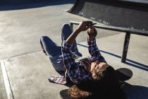 Donna caucasica sdraiata sulla scala con skateboard, utilizzando smartphone al sole. uscire in estate in uno skatepark urbano. — Foto stock