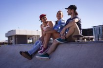 Drei glückliche kaukasische Freundinnen und Freunde sitzen an der Wand und lachen in der Sonne. Im Sommer im städtischen Skatepark abhängen. — Stockfoto
