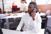 Африканський американець розмовляє на смартфоні, сидячи на столі в сучасному офісі. бізнес, професіоналізм і офісна концепція — стокове фото