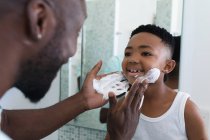 Afro-americano pai e filho no banheiro, aplicando espuma de barbear. em casa em isolamento durante o confinamento de quarentena. — Fotografia de Stock