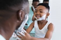 Afro-americanos padre e hijo en el baño, aplicando espuma de afeitar. en casa en aislamiento durante el bloqueo de cuarentena. - foto de stock
