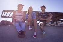 Three happy caucasian female and male friends sitting on wall and laughing in the sun. hanging out at an urban skatepark in summer. — Stock Photo