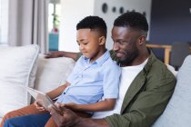 African american father and son sitting on sofa, using tablet and smiling at home in isolation during quarantine lockdown. — Stock Photo