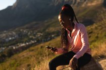 African american woman taking break in exercise outdoors, wearing earphones and using smartphone. healthy active lifestyle and outdoor fitness. — Stock Photo