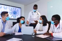 Команда різноманітних лікарів, які носять маску для обличчя, обговорює разом ноутбук у залі засідань. охорона здоров'я та медичні дослідження під час прихованої пандемії 19 — стокове фото