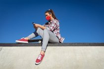Белая женщина в маске, сидит на стене со скейтбордом и пользуется смартфоном. тусоваться в городском скейтпарке летом во время пандемии коронавируса ковид 19. — стоковое фото