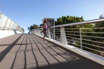 Fit african american woman in face mask stretching on foot bridge exercising in city. healthy active lifestyle and outdoor fitness during coronavirus covid 19 pandemic. — Stock Photo