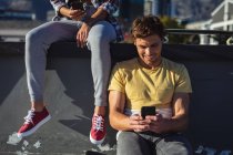 Femme et homme caucasiens assis sur le mur avec des planches à roulettes, en utilisant un smartphone au soleil. traîner dans un skatepark urbain en été. — Photo de stock