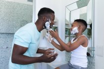Afrikanisch-amerikanischer Vater und Sohn im Badezimmer und tragen Rasierschaum auf. Zuhause in Isolation während der Quarantäne. — Stockfoto