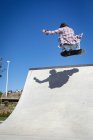 Vista trasera del hombre caucásico saltando y patinando en un día soleado. pasando el rato en skatepark urbano en verano. - foto de stock