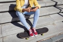 Низька частина білої жінки сидить на сходах зі скейтбордом і використовує смартфон. висить у міському скейтпарку влітку . — стокове фото