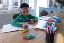 Niño afroamericano en la clase de la escuela en línea, usando el ordenador portátil y la escritura en su cuaderno. en casa en aislamiento durante el bloqueo de cuarentena. - foto de stock