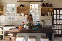 Sorridente donna caucasica che tende a piante in vaso in piedi in cucina cottage soleggiata. vita sana, vicino alla natura fuori dalla griglia casa rurale. — Foto stock