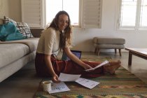 Retrato de mulher caucasiana trabalhando em casa sentado no chão com papelada e laptop sorrindo. trabalhar em casa de forma isolada durante o confinamento de quarentena. — Fotografia de Stock