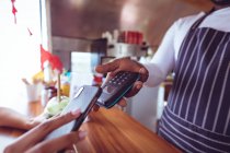 Midsection of african american man in food truck taking smartphone payment holding terminal. independent business and street food service concept. — Stock Photo