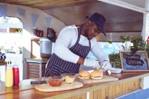 Afrikanisch-amerikanischer Mann im Foodtruck bereitet Ordnung mit Hamburgern auf Arbeitsplatte. Unabhängiges Geschäfts- und Streetfood-Konzept. — Stockfoto