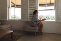 Mulher caucasiana feliz usando fones de ouvido usando smartphone sentado à janela na sala de estar cottage. simples viver em uma casa rural fora da grade. — Fotografia de Stock