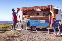 Vielfältiges Paar öffnet und bereitet am sonnigen Tag einen Foodtruck am Meer zu. Unabhängiges Geschäfts- und Streetfood-Konzept. — Stockfoto