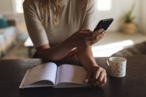 Parte central da mulher sentada na mesa com livro e café usando smartphone. trabalhar em casa de forma isolada durante o confinamento de quarentena. — Fotografia de Stock