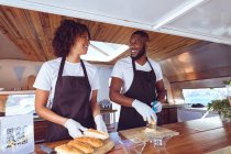 Smiling diverse couple preparing hot dogs behind counter in food truck. independent business and street food service concept. — Stock Photo