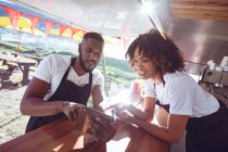 Smiling diverse couple leaning on counter using tablet in food truck. independent business and street food service concept. — Stock Photo