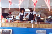 Diverse couple preparing hot dogs behind counter in food truck. independent business and street food service concept. — Stock Photo