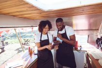 Smiling diverse couple behind counter using tablet in food truck. independent business and street food service concept. — Stock Photo
