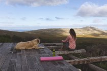 Mujer caucásica pensativa sentada en la cubierta con perro mascota admirando la vista en el entorno rural de montaña. vida sana, fuera de la red y cerca de la naturaleza. - foto de stock