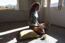 Happy caucasian woman sitting on floor using laptop in sunny cottage living room. simple living in an off the grid rural home. — Stock Photo