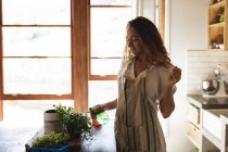 Femme caucasienne souriante tendant aux plantes en pot debout dans la cuisine de chalet ensoleillée. mode de vie sain, proche de la nature dans la maison rurale hors réseau. — Photo de stock