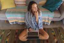 Mulher caucasiana cuidadosa com cobertor sentado no chão usando laptop na sala de estar ensolarada casa de campo. simples viver em uma casa rural fora da grade. — Fotografia de Stock