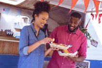 Smiling diverse couple eating hot dogs and potato wedges by food truck on sunny day. independent business and street food service concept. — Stock Photo