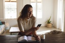Femme blanche assise au bureau avec livre et café à l'aide d'un smartphone. travailler à domicile en isolement pendant le confinement en quarantaine. — Photo de stock