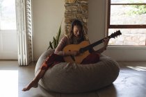 Mujer caucásica feliz sentado en beanbag tocando la guitarra acústica en la sala de estar casa soleada. simple vivir en una casa rural fuera de la red. - foto de stock
