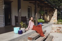 Smiling caucasian woman sitting on steps relaxing on sunny cottage terrace. healthy living, close to nature in off the grid rural home. — Stock Photo