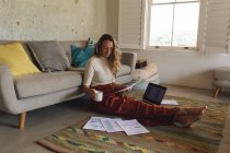 Femme blanche travaillant à la maison assis sur le sol avec de la paperasse et ordinateur portable tenant du café. travailler à domicile en isolement pendant le confinement en quarantaine. — Photo de stock