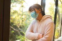 Triste menina asiática em óculos usando máscara facial e olhando para fora da janela. em casa, isoladamente, durante o bloqueio da pandemia e quarentena do covid 19. — Fotografia de Stock
