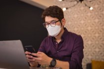 Азиатский бизнесмен в маске для лица использует смартфон и ноутбук в лобби отеля. Цифровой кочевник в городе во время ковида 19 пандемической концепции. — стоковое фото