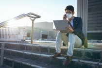 Азиатский бизнесмен в маске с помощью смартфона и ноутбука сидит на ступеньках на городской улице. Цифровой кочевник в городе во время ковида 19 пандемической концепции. — стоковое фото
