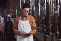 Caucasian male worker wearing apron writing on clipboard at gin distillery. alcohol production and filtration concept — Stock Photo