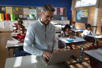 Insegnante maschio caucasico che usa il computer portatile mentre è seduto nella classe alla scuola elementare. concetto di scuola e istruzione — Foto stock