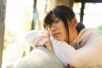 Retrato de niña asiática reflexiva en gafas sentadas en el sofá. en casa en aislamiento durante el bloqueo de cuarentena. - foto de stock