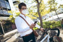 Asian businessman wearing face mask using smartphone holding bike in city street. digital nomad out and about in city during covid 19 pandemic concept. — Stock Photo