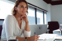 Lächelnde kaukasische Geschäftsfrau sitzt am Schreibtisch und macht sich Notizen bei der Arbeit. unabhängiges kreatives Geschäft in einem modernen Büro. — Stockfoto