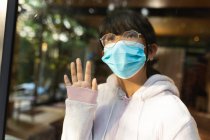 Грустная азиатка в очках в маске и выглядывает в окно. в доме в изоляции во время ковида 19 пандемии и карантинной изоляции. — стоковое фото