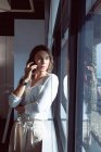 Mujer de negocios caucásica de pie en la ventana, hablando por teléfono inteligente en el trabajo. negocio creativo independiente en una oficina moderna - foto de stock