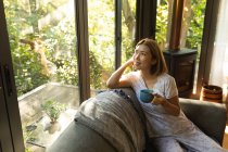 Портрет улыбающейся азиатки, держащей чашку чая и сидящей на диване. в доме в изоляции во время карантинной изоляции. — стоковое фото