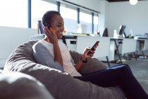 Donna d'affari afroamericana sorridente seduta in poltrona, con le cuffie, usando lo smartphone. attività creativa indipendente in un ufficio moderno. — Foto stock